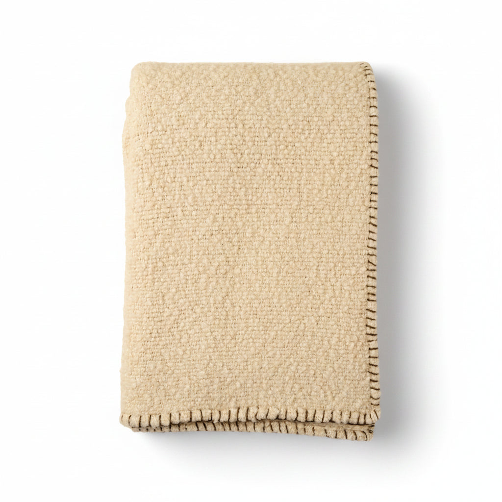 A folded cream-colored textured throw blanket with a dark brown fringed edge, displayed on a solid cream background.