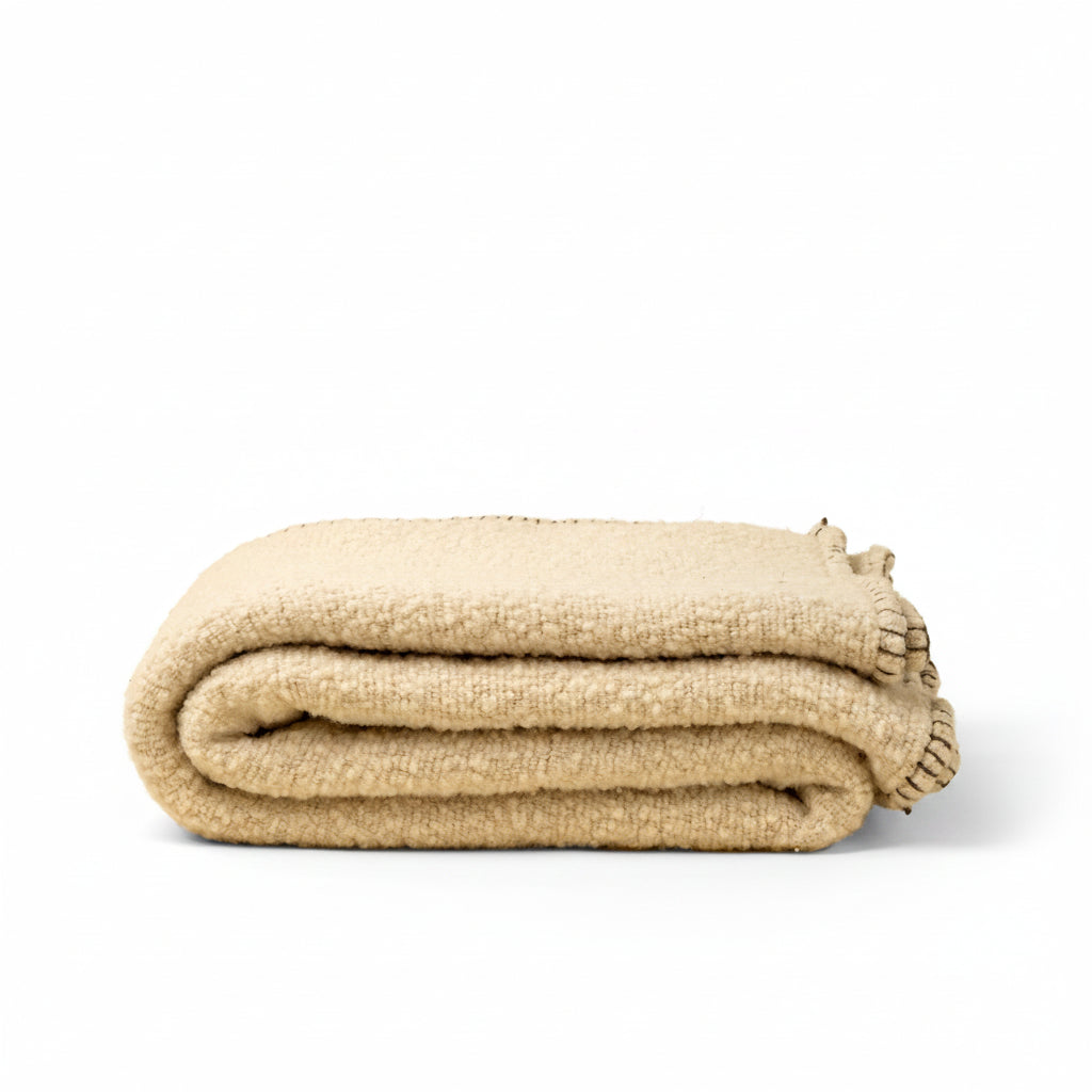 A folded beige textured blanket with fringed edges, photographed on a solid cream background.