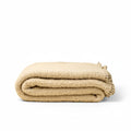 A folded beige textured blanket with fringed edges, photographed on a solid cream background.
