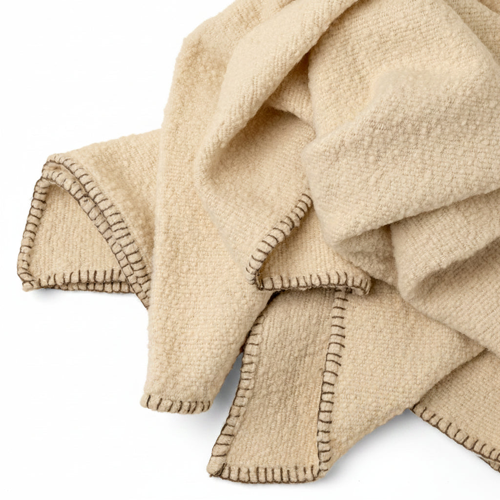 A close-up of a soft woven throw blanket in a warm cream or oatmeal tone, draped to show its textured surface and contrast whipstitch trim on a solid light beige background.